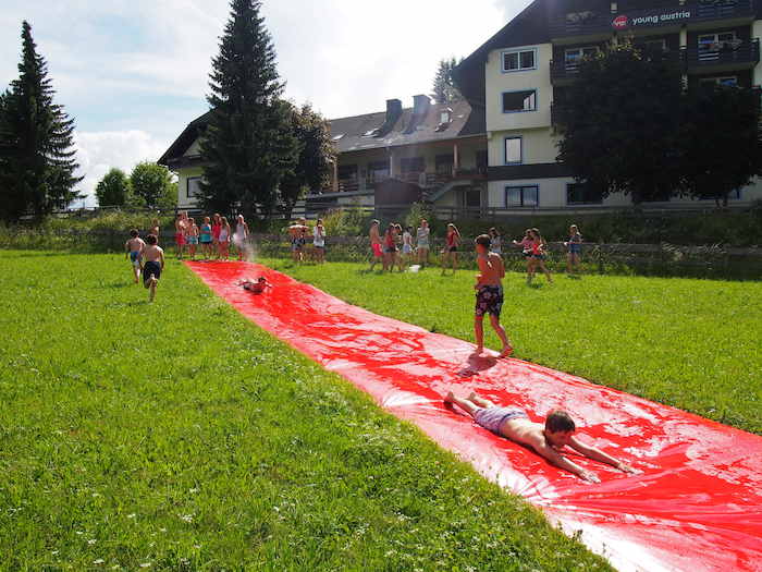 Al campo avventura in Austria due ragazzi scivolano a pancia in giù da un tappeto d'acqua rosso messo sul prato in pendenza