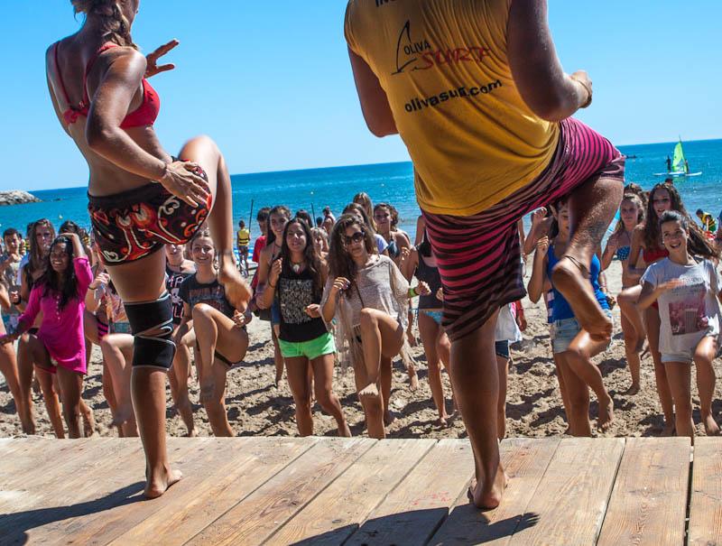 In questo campo estivo surf tanti ragazzi di tutte le età ballano in gruppo in riva al mare di fronte a due animatori che fanno vedere le mosse da fare