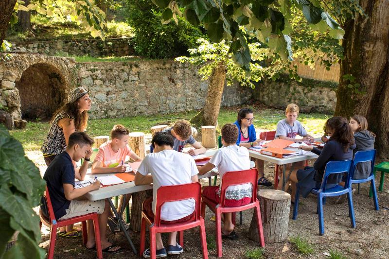 I ragazzi del soggiorno linguistico in francese sono seduti ad un tavolo all'aperto e stanno studiando mentre un'istruttrice li aiuta da in piedi