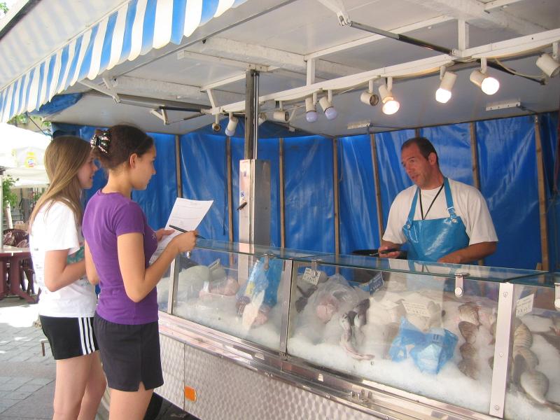 Due ragazze del campo estivo Germania sono al mercato davanti al bancone del pesce e stanno parlando con il venditore usando i loro appunti