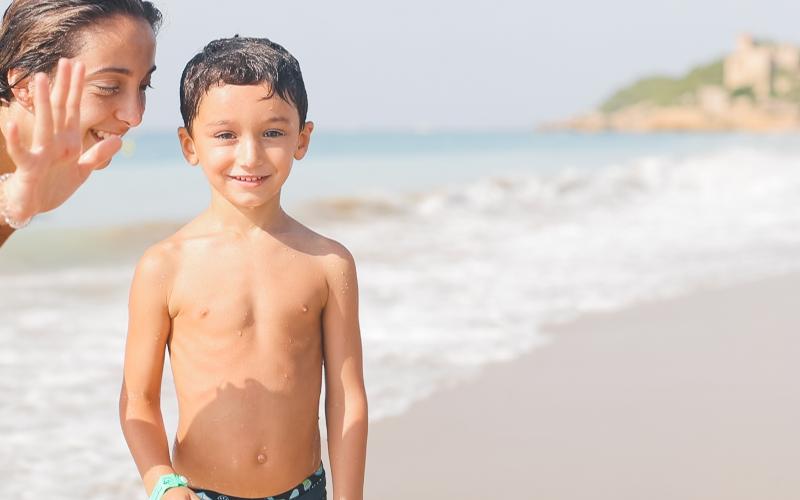 Un bambino in costume sorride in riva al mare al campo estivo attività in Spagna mentre una ragazza lo guarda e saluta