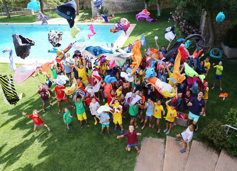 Presso il campo estivo in Spagna, tutti i ragazzi festeggiano a bordo piscina lanciando in aria palloncini e giochi