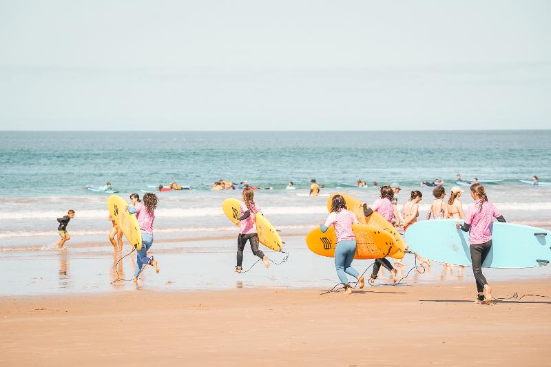 I partecipanti del campo estivo surf corrono sulla spiaggia verso il mare con le tavole sotto braccio