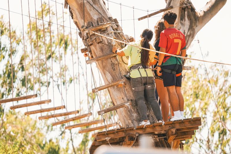 Tre partecipanti del campo estivo surf aspettano il loro turno per attraversare il percorso sospeso tra gli alberi