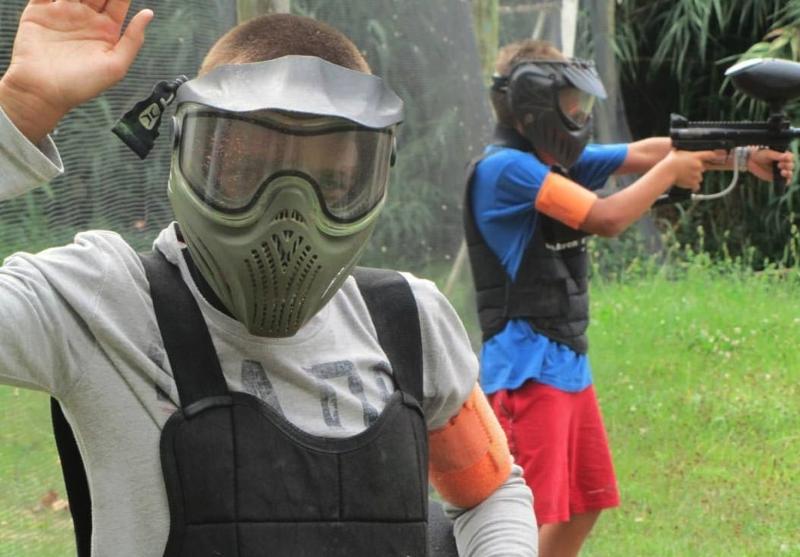 Un ragazzo che indossa un casco protettivo saluta la fotocamera mentre il suo amico sta sparando agli avversari durante la partita di Paintball del soggiorno linguistico Germania