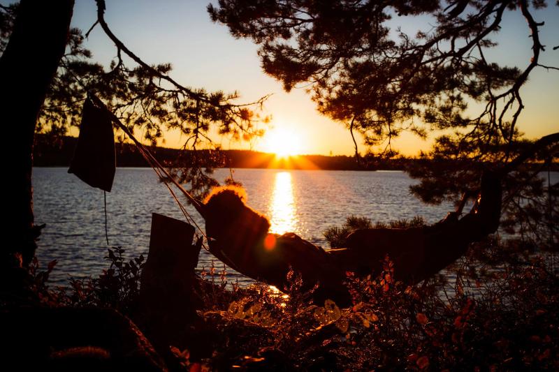 Un partecipante del campo avventura in Svezia è sdraiato in un'amaca e guarda il tramonto all'orizzonte che si riflette sull'acqua del lago