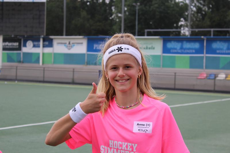 Una ragazza vestita di rosa con una fascia e un polsino bianchi sorride e fa il pollice alzato al campo estivo internazionale di Hockey a Rotterdam