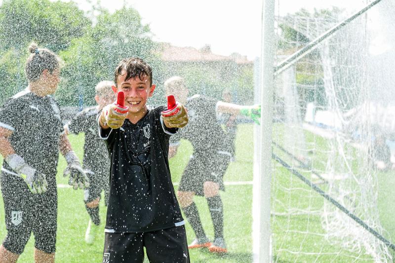 Un giovane portiere del campus estivo calcio sorride divertito sotto l'acqua degli irrigatori dei campi