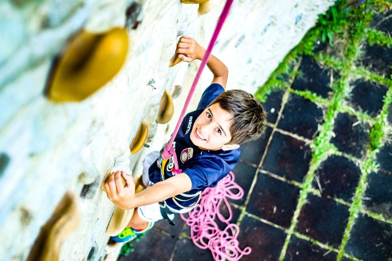  Na imagem, um rapaz jovem, sorridente e equipado com arnês, sobe uma parede de escalada, concentrado e entusiasmado com o desafio, representando a experiência aventureira e ativa que um campo de férias itália oferece.