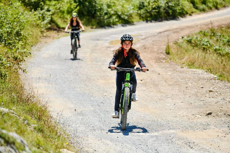  Na imagem, duas jovens andam de bicicleta por um trilho de terra rodeado por vegetação, com capacetes e sorrisos que refletem a diversão e aventura de explorar o campo de férias itália em duas rodas.