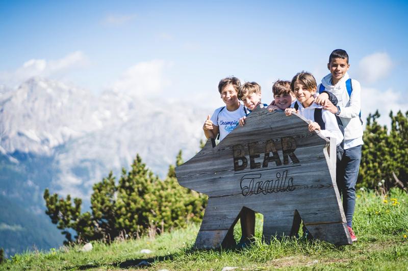 Alcuni partecipanti del campo estivo Padel sono appoggiati su una statua di legno rappresentante un orso e danno le spalle alle montagne