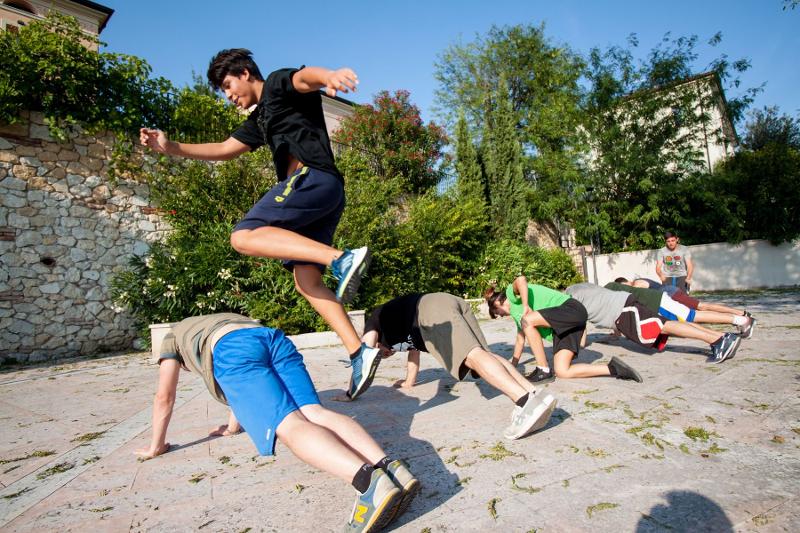 Un ragazzo del campo estivo Parkour salta agilmente i suoi compagni mentre fanno dei piegamenti sulle braccia