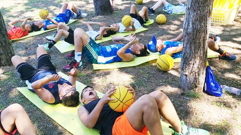 Bambini sdraiati per terra con materassini si allenano con palle da basket al campo estivo All Star basket a Lignano Sabbiadoro